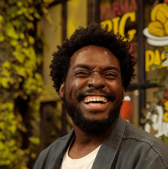 Close-up, circular photo of a young man with a warm smile, short dark curly hair, and a dark beard, looking directly at the viewer.